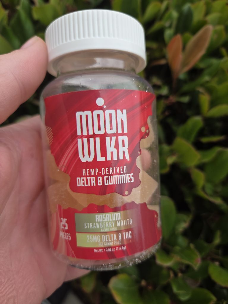 MOONWLKR Delta 8 Gummies — Strawberry Mojito container with red gummies inside being held by a person on top of a bush.