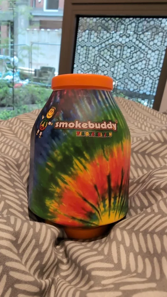 Personal air filter for dabs from Smokebuddy  displayed on a bed with an overlooking window showing the outside trees.