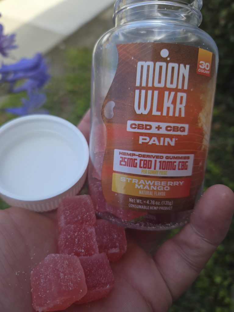 MOONWLKR CBD:CBG Pain Relief Gummies held by a person, showing the red gummies, the jar cover, and the jar itself, outdoors.