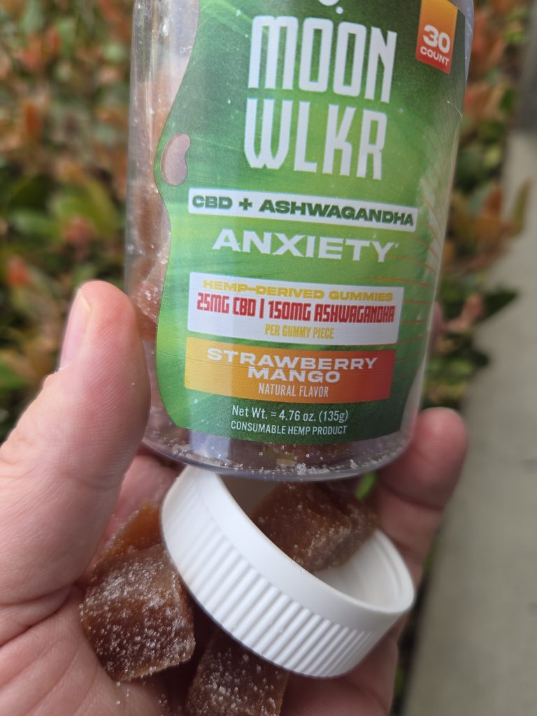 MOONWLKR CBD:Ashwagandha Stress Relief Gummies — Strawberry Mango container being held by a person showing the cover and one piece of gummy.