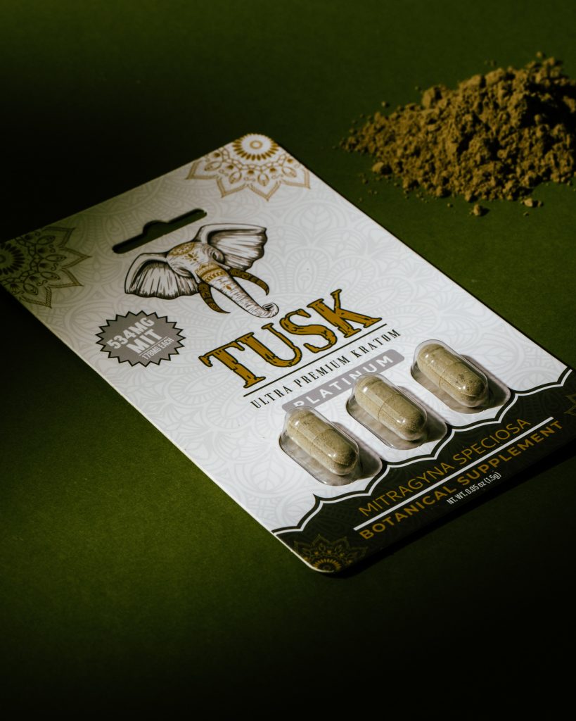 Kratom capsule inside its Tusk Kratom packaging displayed on top of a green sheet with green kratom powder piled up beside it.