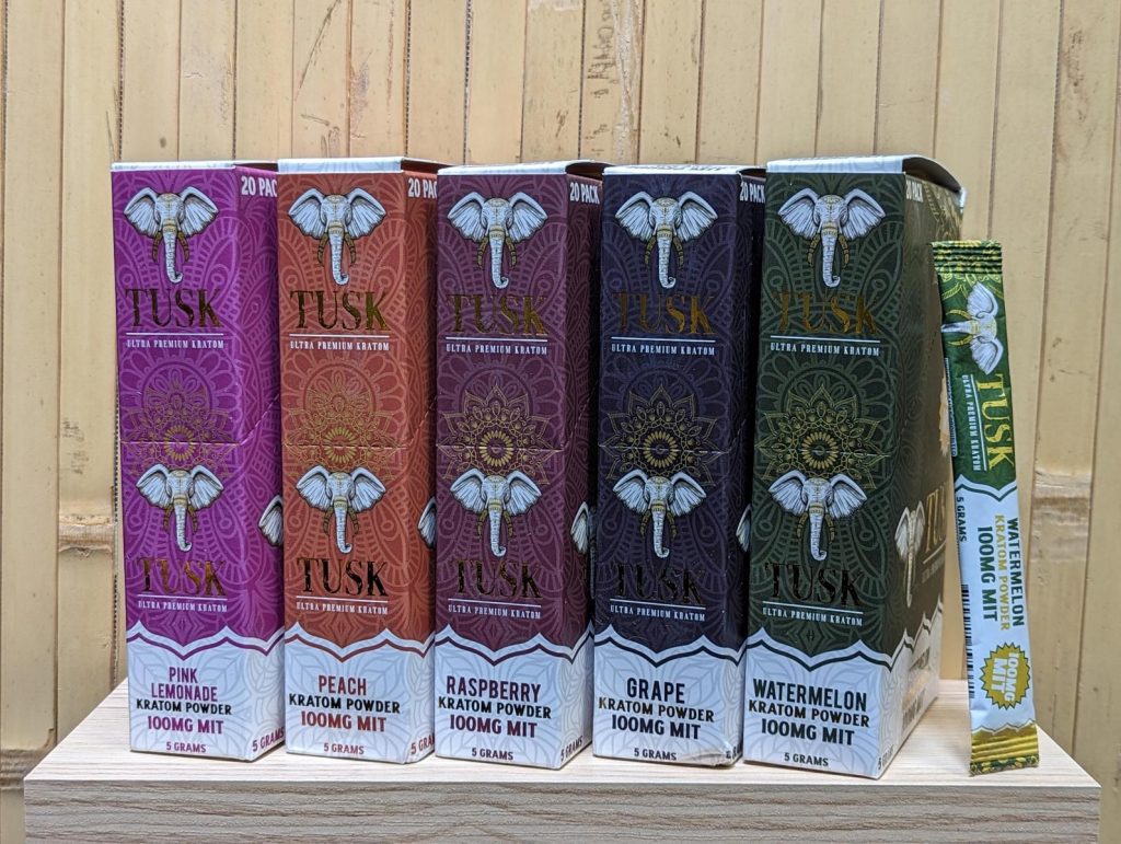 Single-serve kratom products from Tusk Kratom aligned side-by-side in front of a wooden background outdoors.