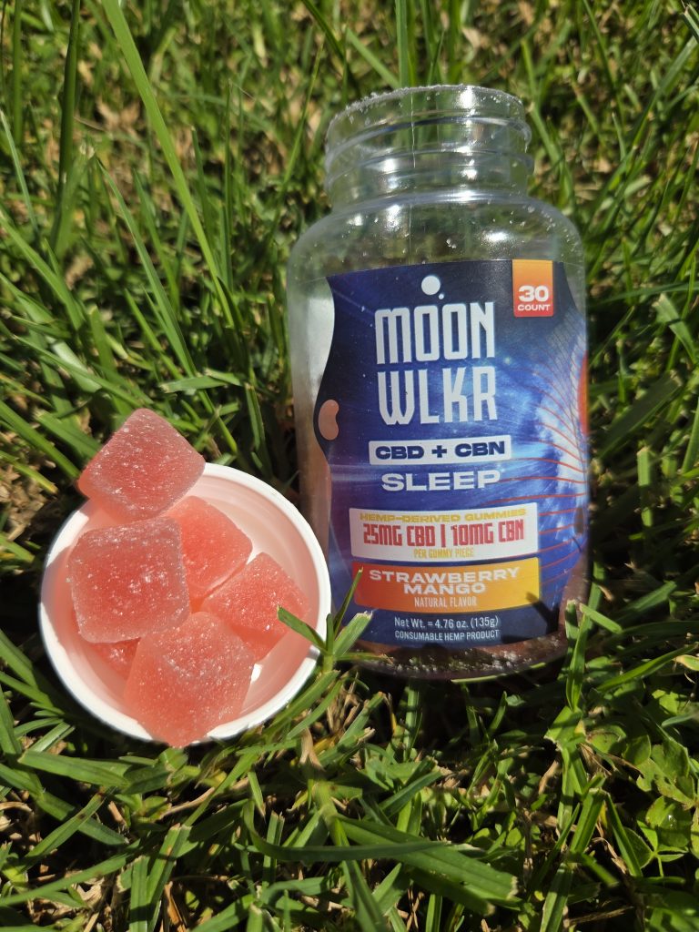 MOONWLKR CBD:CBN Sleep Gummies — Strawberry Mango displayed on a patch of grass showing the jar and the gummy.